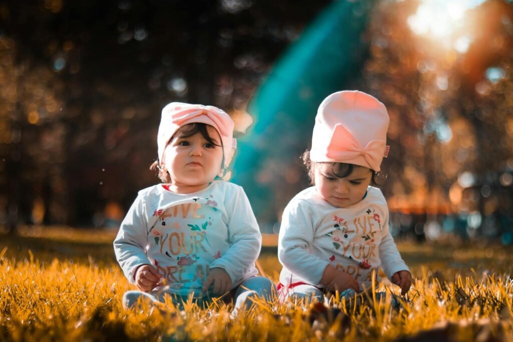 Charming twin babies in matching outfits play on grass under autumn sun. Perfect fall moment captured.